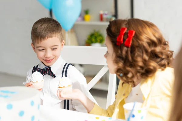 Adorables niños sentados en la mesa y sosteniendo cupcakes durante la fiesta de cumpleaños en casa - foto de stock