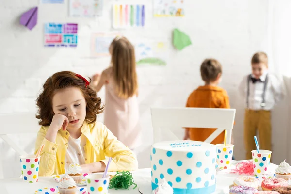 Selective focus of sad kid sitting at table with cake during birthday party — Stock Photo