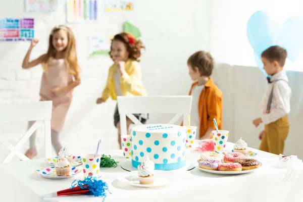 Niños divirtiéndose durante la fiesta de cumpleaños cerca de la mesa con pastel, rosquillas y cupcakes en casa - foto de stock