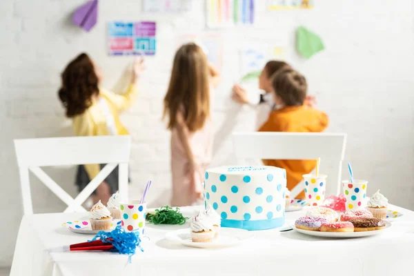 Enfoque selectivo de la mesa con pastel de cumpleaños, cuernos de fiesta, vasos de papel, magdalenas y rosquillas - foto de stock