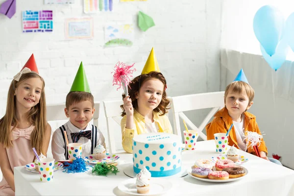 Kinder in Partyhüten sitzen am Tisch und schauen bei Geburtstagsfeier in die Kamera — Stockfoto