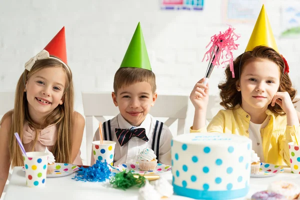 Kinder in Partyhüten sitzen am Tisch und schauen bei Geburtstagsfeier in die Kamera — Stockfoto