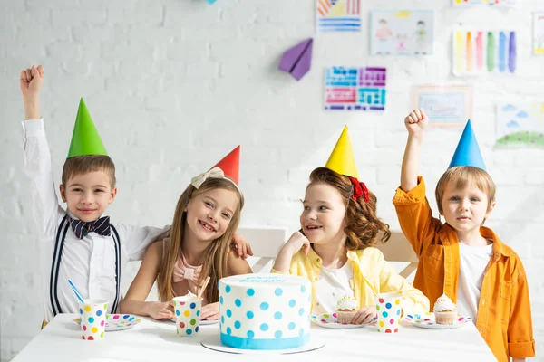Niños felices en sombreros de fiesta sentados en la mesa con pastel y celebrando cumpleaños juntos - foto de stock