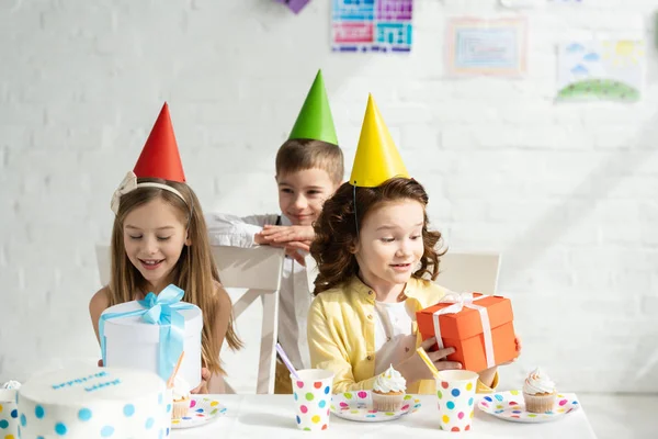 Crianças adoráveis em bonés de festa sentado à mesa com caixas de presente durante a festa de aniversário em casa — Fotografia de Stock