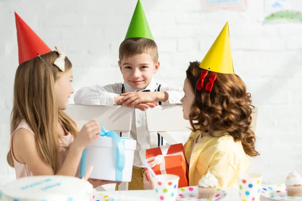 Adorable kids in party caps sitting at table with gift boxes during birthday party at home — Stock Photo