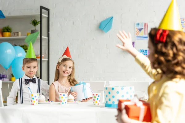 Kid waving to friends in party caps sitting at table during birthday celebration at home — Stock Photo