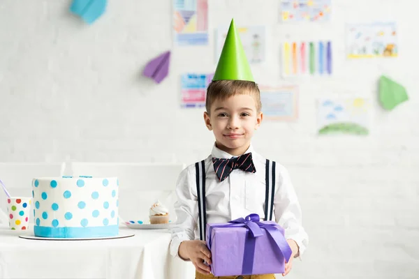 Очаровательный мальчик в шапочке для вечеринок смотрит в камеру и держит подарок во время празднования дня рождения — стоковое фото