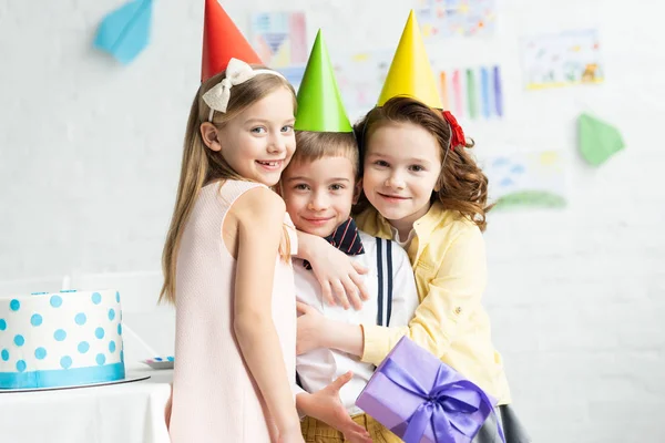 Crianças sorridentes em bonés de festa abraçando menino adorável com caixa de presente durante a festa de aniversário em casa — Fotografia de Stock