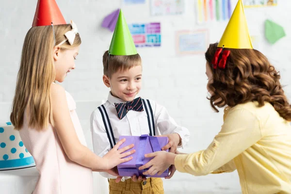 Niños en gorras de fiesta dando caja de regalo para niño durante la fiesta de cumpleaños en casa - foto de stock