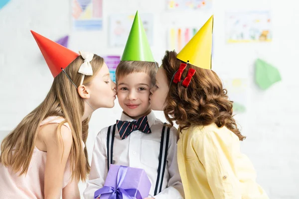Enfants dans les casquettes de fête baisers sur les joues adorable garçon avec boîte cadeau pendant la fête d'anniversaire à la maison — Photo de stock