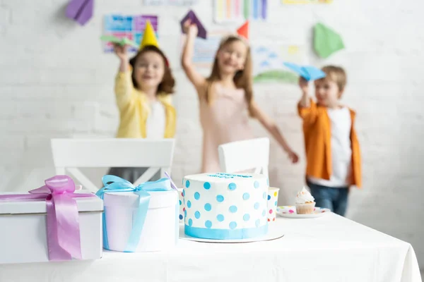 Selective focus of cake and presents on table and kids playing with paper planes during birthday party — Stock Photo