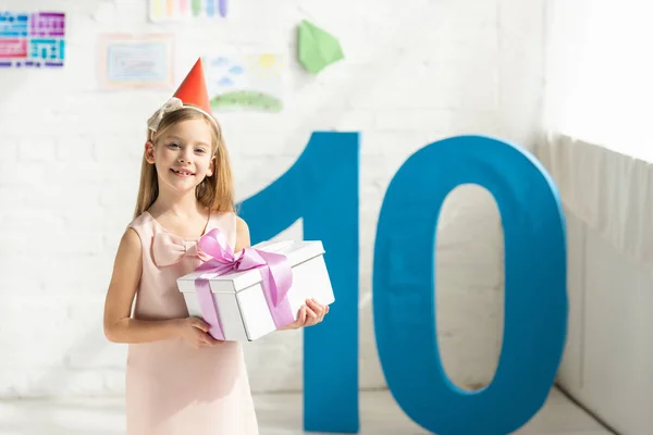 Adorable feliz niño en partido gorra celebración presente y posando cerca de número decorativo 10 - foto de stock