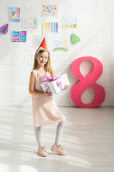 Adorable smiling kid in party cap holding present and posing near decorative pink number 8 — Stock Photo