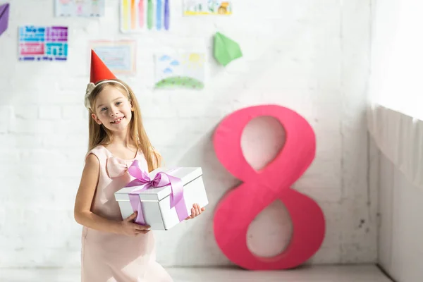 Adorable smiling kid in party cap holding present and posing near decorative pink number 8 — Stock Photo