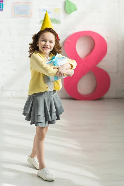 Criança feliz adorável no tampão do partido que prende presente e posando perto do número rosa decorativo 8 — Fotografia de Stock