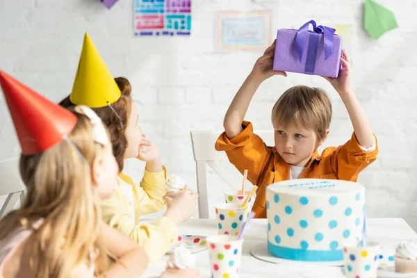 Niño sosteniendo presente mientras está sentado en la mesa con amigos durante la celebración de la fiesta de cumpleaños - foto de stock
