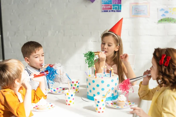 Kids sitting at table with cake and cheering with party horns during birthday celebration — Stock Photo