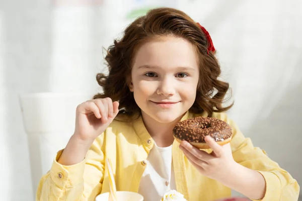 Очаровательный улыбающийся ребенок в желтом держит вкусный пончик и смотрит в камеру — стоковое фото