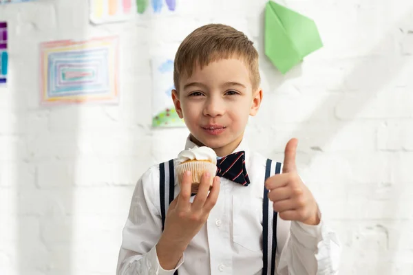Adorável preteen menino no arco gravata segurando cupcake e mostrando polegar para cima sinal — Fotografia de Stock