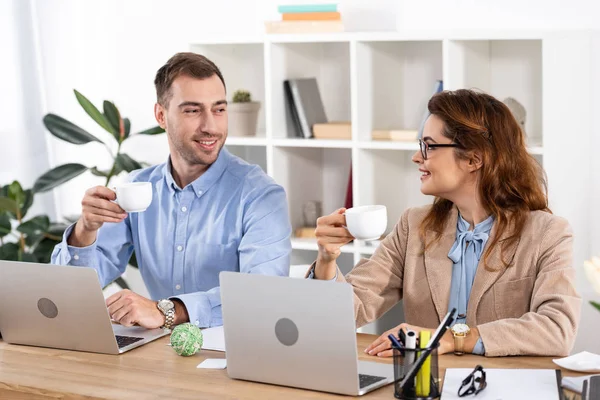 Cheerful businesswoman in glasses holding cup and looking at coworker in office — Stock Photo