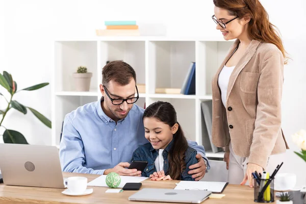 Padre alegre usando teléfono inteligente cerca de hija feliz en la oficina - foto de stock