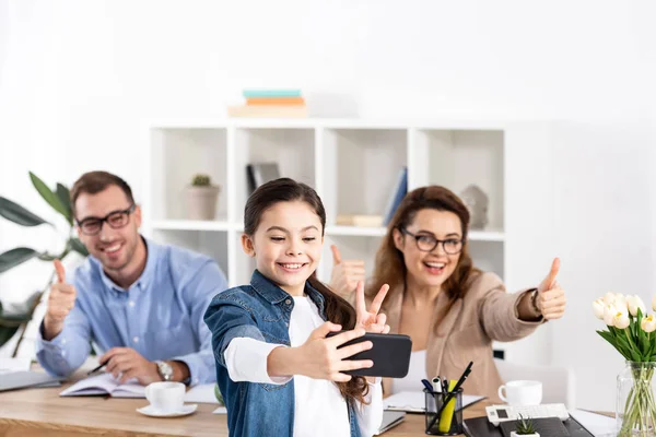 Selective focus of cheerful child taking selfie on smartphone near happy parents gesturing in office — Stock Photo