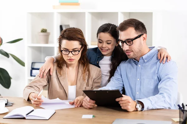 Cheerful kid hugging mother and father in glasses looking at papers in office — Stock Photo