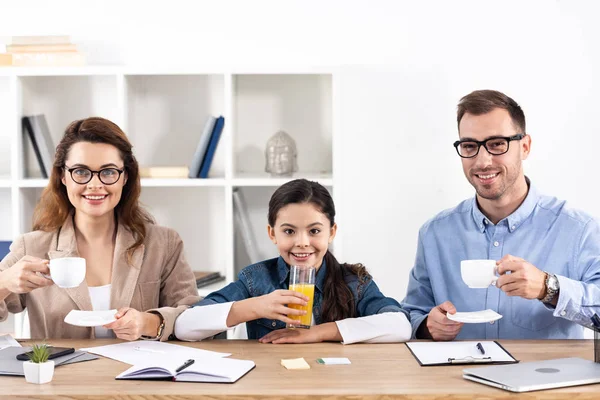 Alegre hombre y mujer sosteniendo tazas cerca de niño con vaso de jugo de naranja - foto de stock