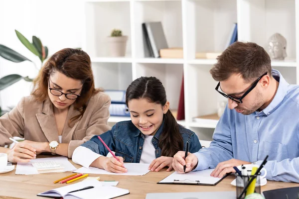 Happy child drawing near parents in glasses working in office — Stock Photo