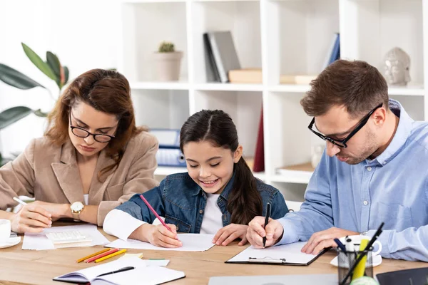 Niño feliz dibujo cerca de los padres en gafas de trabajo en la oficina - foto de stock