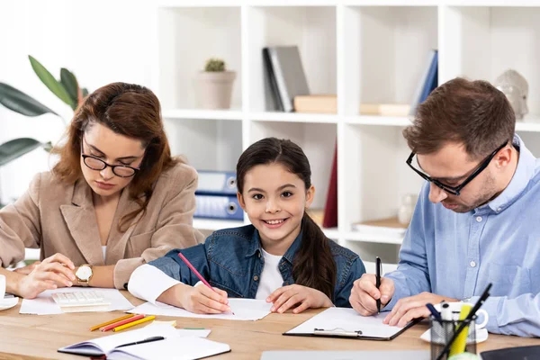 Mignon enfant dessin près des parents dans des lunettes de travail dans le bureau — Photo de stock