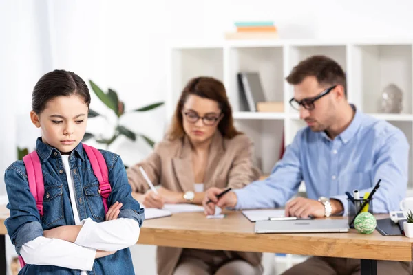 Messa a fuoco selettiva di bambino sconvolto con zaino in piedi con braccia incrociate vicino ai genitori in ufficio — Foto stock