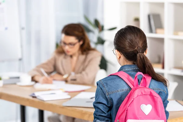 Vista trasera del niño con mochila rosa de pie cerca de la madre que trabaja en la oficina - foto de stock