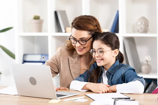 Alegre madre sentado con linda hija en gafas y mirando a la computadora portátil - foto de stock