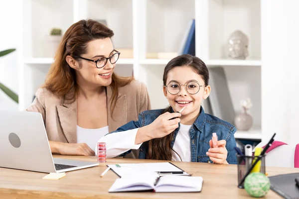 Fröhliche Mutter schaut süße Tochter beim Auftragen von Lipgloss im Büro an — Stockfoto