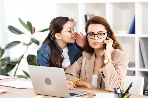 Lindo niño susurrando al oído de la madre hablando en el teléfono inteligente cerca de la computadora portátil en la oficina - foto de stock