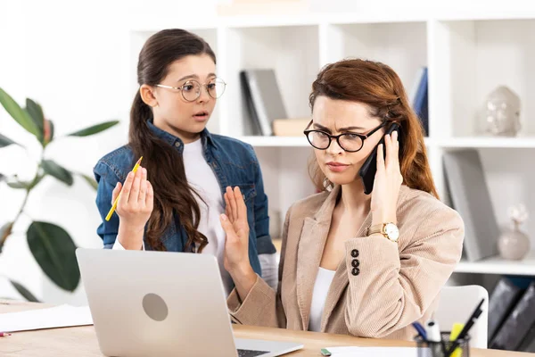 Surprised kid looking at mother talking on smartphone and gesturing in office — Stock Photo