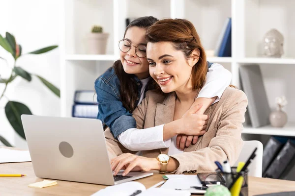 Glückliche Tochter umarmt fröhliche Mutter mit Laptop im Büro — Stockfoto