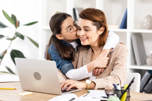 Mignon enfant étreignant et embrassant joue de mère heureuse au bureau — Photo de stock