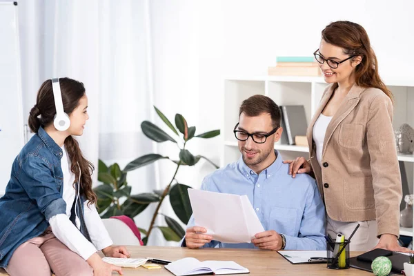 Happy kid listening music in headphones near father holding empty papers in office — Stock Photo