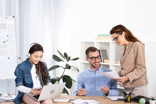 Cheerful kid using laptop while listening music in headphones near parents in office — Stock Photo