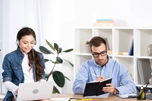 Cheerful kid using laptop while listening music in headphones near father in office — Stock Photo