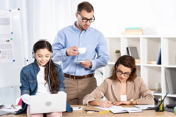 Carino bambino utilizzando il computer portatile durante l'ascolto di musica in cuffie vicino a madre e padre in ufficio — Foto stock