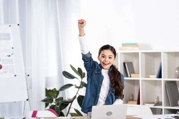 Счастливый ребенок празднует триумф возле ноутбука в современном офисе — стоковое фото