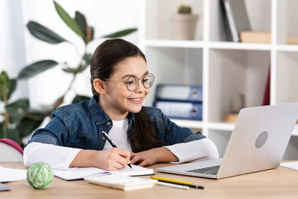 Щаслива дитина дивиться на ноутбук, тримаючи ручку біля блокнота в офісі — стокове фото