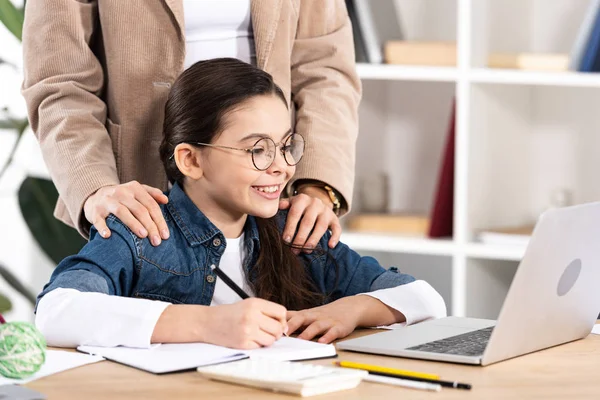 Abgeschnittene Ansicht der Mutter, die Hände auf die Schultern eines glücklichen Kindes legt, das im Büro auf den Laptop schaut — Stockfoto