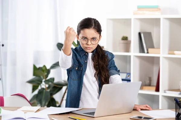 Mignon enfant dans des lunettes montrant poing tandis que debout près de l'ordinateur portable dans le bureau — Photo de stock