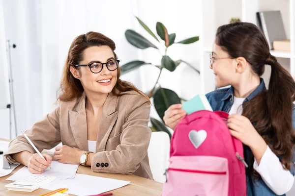 Fröhliche Mutter mit Brille schaut süße Tochter mit Rucksack im Büro an — Stockfoto