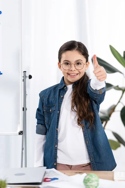 Весёлый ребенок, показывающий большой палец возле белой доски в офисе — стоковое фото