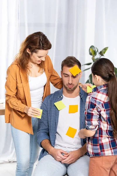 Happy woman and kid putting yellow sticky notes on upset man — Stock Photo